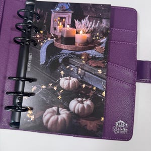 Puede incluir: Una agenda morada con anillas negras, abierta a un inserto decorativo. El inserto presenta una escena oto&ntilde;al con calabazas, velas y el texto "Breathe & Budget". La agenda tiene ranuras para tarjetas y un cierre a presi&oacute;n.