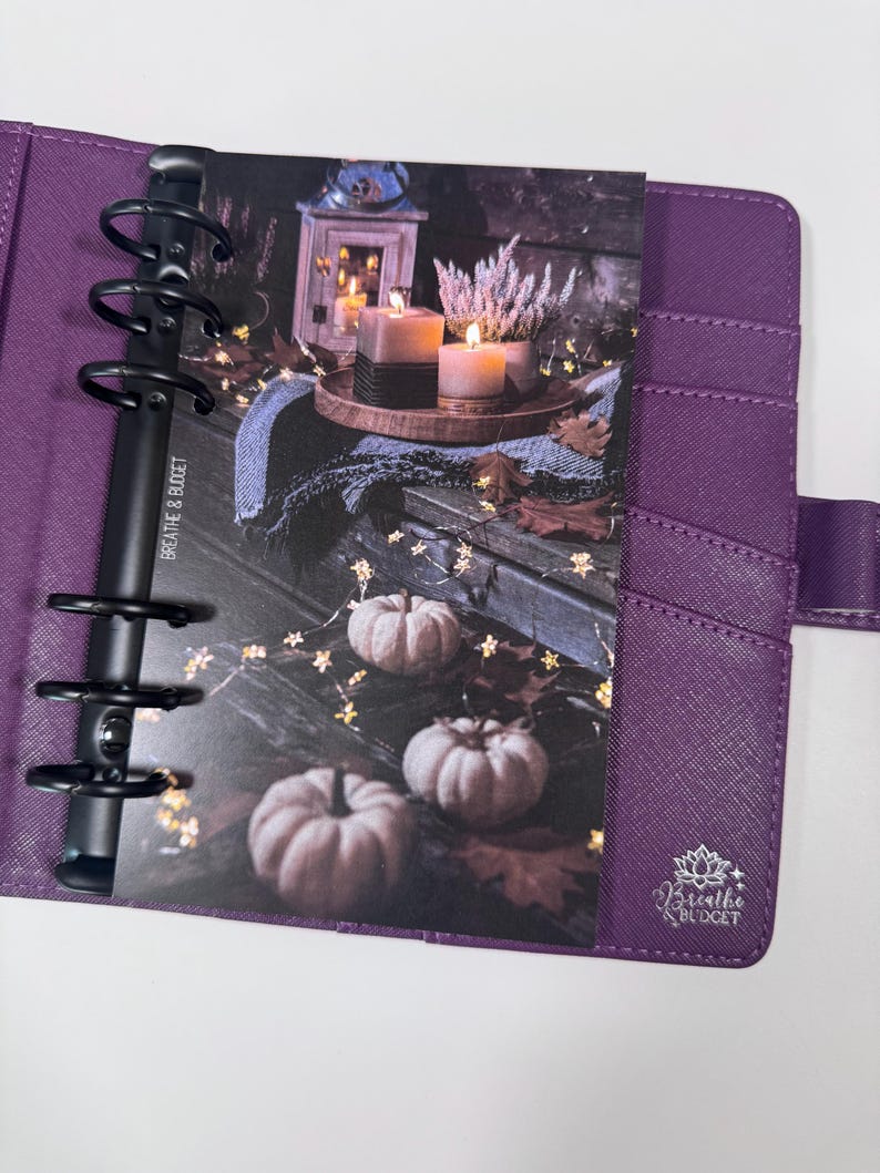 Puede incluir: Una agenda morada con anillas negras, abierta a un inserto decorativo. El inserto presenta una escena oto&ntilde;al con calabazas, velas y el texto "Breathe & Budget". La agenda tiene ranuras para tarjetas y un cierre a presi&oacute;n.