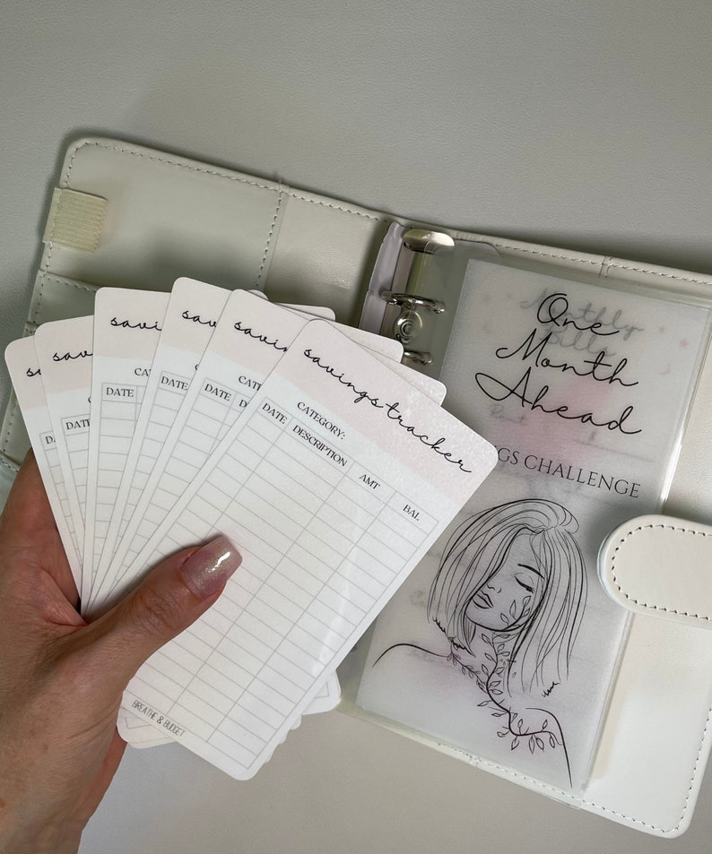 Op de afbeelding: Een witte map met een doorzichtig plastic zakje dat een spaar-trackerblad bevat met de tekst "One Month Ahead Savings Challenge". Het blad toont een lijntekening van een vrouw met bladeren in haar haar. De map bevat ook verschillende witte kaarten met de tekst "savings tracker" en kolommen voor datum, categorie, beschrijving, bedrag en saldo.
