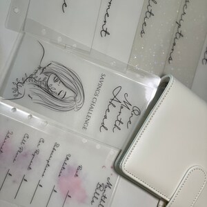 Op de afbeelding: Een witte planner met een doorzichtig plastic vel er bovenop. Het vel heeft een tekening van een vrouwen gezicht met bladeren rond haar hoofd. De tekst op het vel zegt "One Month Ahead" en "Savings Challenge". De planner heeft een sectie voor "Monthly Bills" met categorie&euml;n voor "Rent", "Utilities", "Subscriptions", "Car Payment", en "Cell Phone".