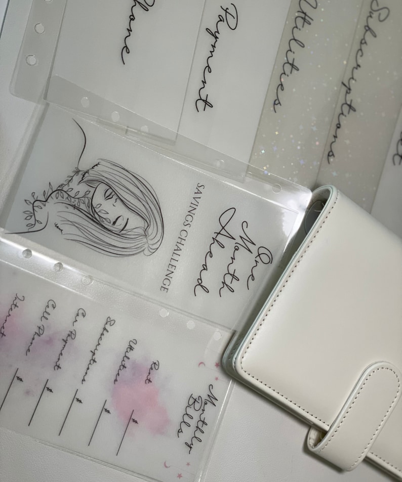 Op de afbeelding: Een witte planner met een doorzichtig plastic vel er bovenop. Het vel heeft een tekening van een vrouwen gezicht met bladeren rond haar hoofd. De tekst op het vel zegt "One Month Ahead" en "Savings Challenge". De planner heeft een sectie voor "Monthly Bills" met categorie&euml;n voor "Rent", "Utilities", "Subscriptions", "Car Payment", en "Cell Phone".