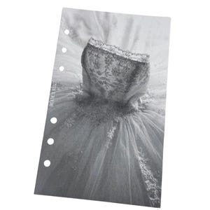 Puede incluir: Una imagen en blanco y negro de un vestido de novia con detalles de encaje. El vestido est&aacute; colocado sobre una superficie blanca con un borde perforado.