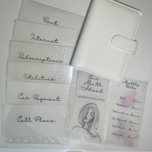 Op de afbeelding: Een witte planner met een doorzichtig plastic zakje aan de voorkant. Het zakje bevat een vel papier met de tekst "One Month Ahead Savings Challenge" en een lijntekening van een vrouw. De planner heeft ook een doorzichtig plastic zakje aan de achterkant met een vel papier waarop de tekst "Monthly Bills" staat en een lijst met rekeningen om bij te houden.