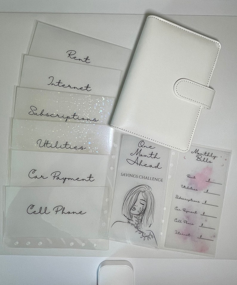 Op de afbeelding: Een witte planner met een doorzichtig plastic zakje aan de voorkant. Het zakje bevat een vel papier met de tekst "One Month Ahead Savings Challenge" en een lijntekening van een vrouw. De planner heeft ook een doorzichtig plastic zakje aan de achterkant met een vel papier waarop de tekst "Monthly Bills" staat en een lijst met rekeningen om bij te houden.