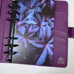 Puede incluir: Agenda morado con anillas met&aacute;licas negras, que contiene una tarjeta con un dise&ntilde;o de hojas azules y moradas. El planificador tiene bolsillos y un cierre de correa. La tarjeta tiene el texto "Breathe & Budget".