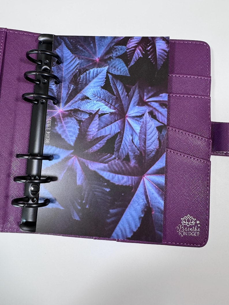 Puede incluir: Agenda morado con anillas met&aacute;licas negras, que contiene una tarjeta con un dise&ntilde;o de hojas azules y moradas. El planificador tiene bolsillos y un cierre de correa. La tarjeta tiene el texto "Breathe & Budget".