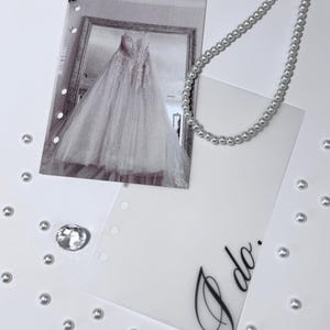 Puede incluir: Una tarjeta blanca con las palabras "Yo s&iacute;" escritas en cursiva negra. La tarjeta est&aacute; rodeada de cuentas de perlas blancas y una foto de un vestido de novia blanco.
