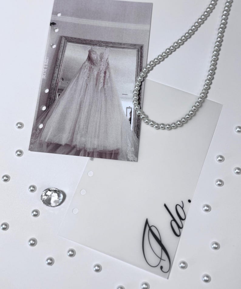 Puede incluir: Una tarjeta blanca con las palabras "Yo s&iacute;" escritas en cursiva negra. La tarjeta est&aacute; rodeada de cuentas de perlas blancas y una foto de un vestido de novia blanco.