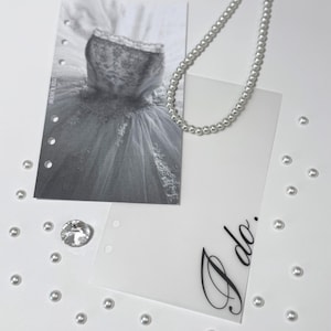 Puede incluir: Una tarjeta blanca con las palabras "I do" escritas en cursiva negra. La tarjeta est&aacute; rodeada de cuentas de perlas blancas y una foto en blanco y negro de un vestido de novia.