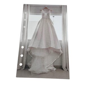 Puede incluir: Un vestido de novia blanco con escote en V y una larga cola cuelga de una percha frente a una ventana.