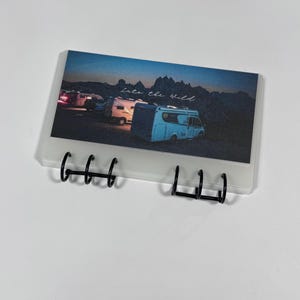 May include: A white, spiral-bound notebook with a clear front cover featuring a photograph of three camper vans parked in front of a mountain range at dusk. The text "Into the Wild" is written in white script on the cover.