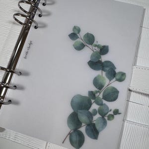 Puede incluir: Una carpeta blanca con una hoja de plástico transparente en el interior. La hoja tiene una ilustración de acuarela de hojas de eucalipto y el texto "breathe & budget".