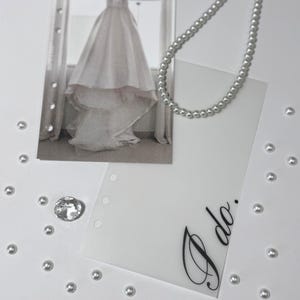 Puede incluir: Una tarjeta blanca con las palabras "S&iacute;, quiero" escritas en cursiva negra. La tarjeta est&aacute; rodeada de perlas blancas y una foto de un vestido de novia blanco.