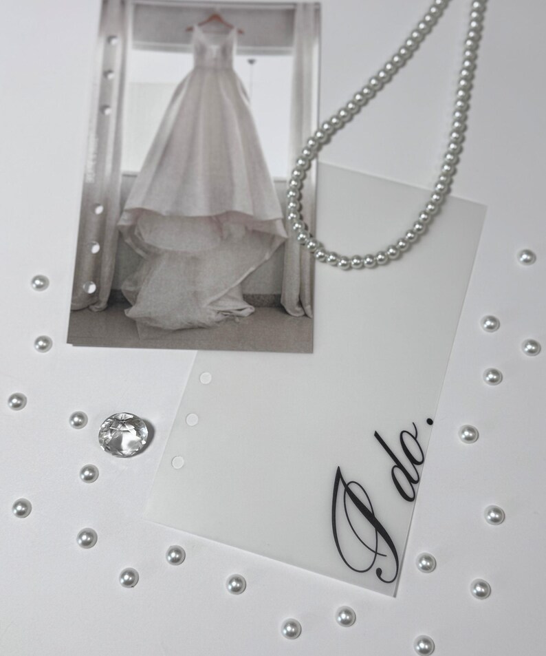 Puede incluir: Una tarjeta blanca con las palabras "S&iacute;, quiero" escritas en cursiva negra. La tarjeta est&aacute; rodeada de perlas blancas y una foto de un vestido de novia blanco.