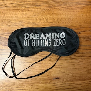 May include: Black sleep mask with the text "DREAMING OF HITTING ZERO" in silver glitter lettering. The mask has a black elastic strap for securing it around the head. The sleep mask is designed to block out light.