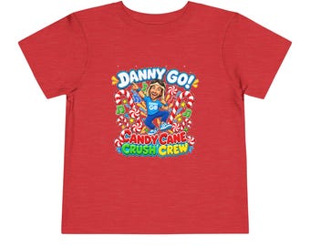 Puede incluir: Una camiseta roja de manga corta con un estampado gráfico de un personaje de dibujos animados con camisa y gorro azules. El texto DANNY GO! CANDY CANE CRUSH CREW aparece impreso sobre y bajo el personaje, rodeado de bastones de caramelo.