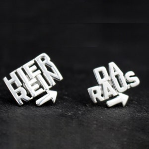 Könnte beinhalten: Ein Paar silberne Ohrringe mit den Worten "Hier Rein" und "Da Raus" darauf. Die Wörter sind so angeordnet, dass sie einen Pfeil bilden, der nach rechts zeigt.