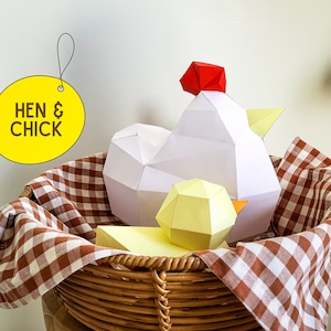 May include: A white paper hen and a yellow paper chick in a brown wicker basket lined with a red and white gingham tablecloth. The basket is labeled "Hen & Chick".