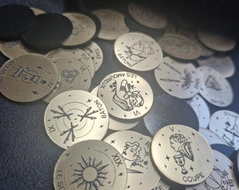May include: A collection of gold-colored tarot card tokens with black etched designs and French text. The tokens feature various illustrations, including a sun, a woman, and symbols. Some tokens are black. The tokens are scattered on a dark surface.