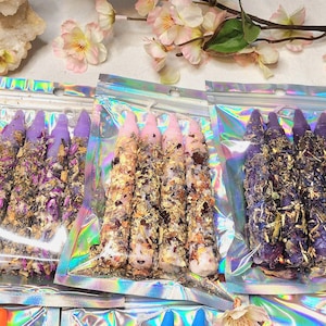 May include: Five holographic pouches each containing three taper candles coated in dried flower petals and herbs. The candles are white, purple, pink, dark purple, and light green, all decorated with botanical elements and encased in clear iridescent plastic.
