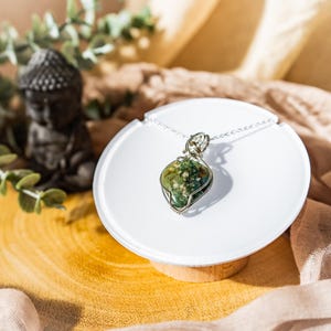 Puede incluir: Un colgante de alambre plateado con una piedra de ágata musgosa verde y marrón. El colgante se muestra en un soporte circular blanco.