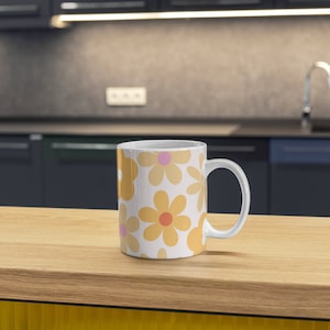 K&ouml;nnte beinhalten: Eine wei&szlig;e Keramiktasse mit einem Blumenmuster. Die Tasse zeigt ein sich wiederholendes Muster aus gelben und orangefarbenen Blumen mit rosa Zentren. Die Tasse steht auf einer Holzoberfl&auml;che.