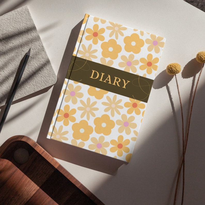 K&ouml;nnte beinhalten: Ein wei&szlig;es Tagebuch mit einem Blumenmuster in Gelb- und Rosat&ouml;nen. Das Wort "DIARY" ist in brauner Schrift auf einem dunkelbraunen Streifen auf dem vorderen Umschlag geschrieben.
