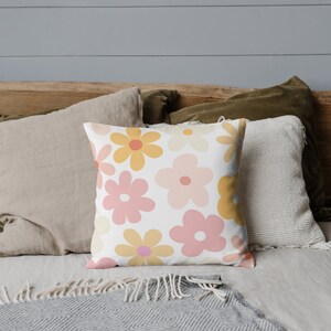 K&ouml;nnte beinhalten: Ein wei&szlig;es Kissen mit einem Retro-Blumenmuster mit rosa, gelben und orangefarbenen Blumen. Das Kissen liegt auf einem Bett mit anderen Kissen und einer Decke.