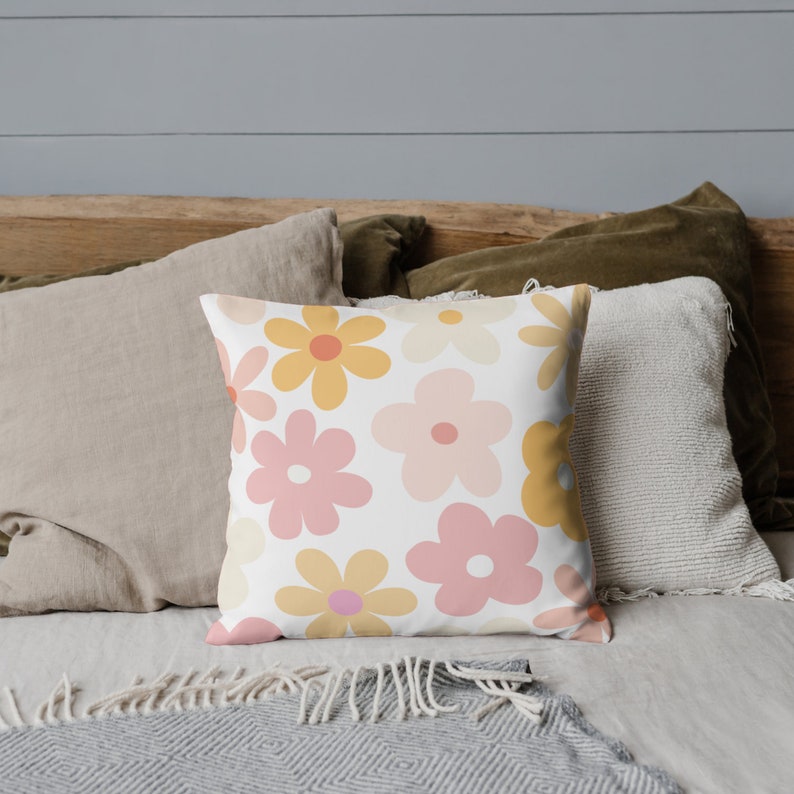 K&ouml;nnte beinhalten: Ein wei&szlig;es Kissen mit einem Retro-Blumenmuster mit rosa, gelben und orangefarbenen Blumen. Das Kissen liegt auf einem Bett mit anderen Kissen und einer Decke.