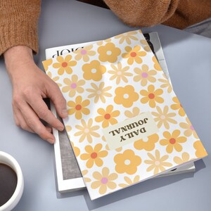 K&ouml;nnte beinhalten: Ein wei&szlig;es Journal mit einem gelben und orangefarbenen Blumenmuster. Das Journal hat den Text "DAILY JOURNAL" auf der Vorderseite.