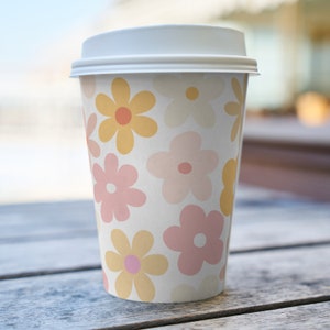 K&ouml;nnte beinhalten: Ein wei&szlig;er To-Go-Becher mit einem Blumenmuster. Das Muster zeigt rosa und gelbe Blumen auf wei&szlig;em Hintergrund.