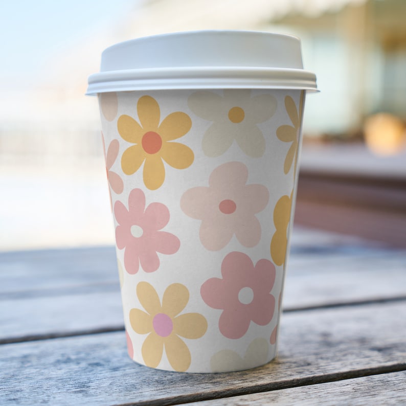 K&ouml;nnte beinhalten: Ein wei&szlig;er To-Go-Becher mit einem Blumenmuster. Das Muster zeigt rosa und gelbe Blumen auf wei&szlig;em Hintergrund.