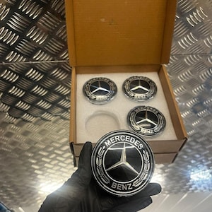 May include: Set of four Mercedes-Benz wheel center caps in a cardboard box. The caps are black and silver with the Mercedes-Benz logo. One cap is held in a gloved hand, showcasing the design. The box is on a metal surface.