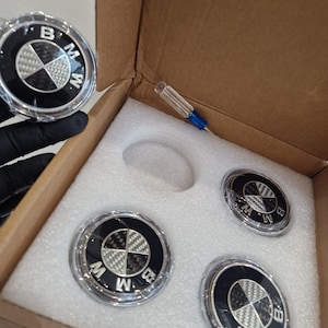 May include: Set of four BMW wheel center caps with a carbon fiber design. The caps feature the BMW logo in silver against a black background. One cap is held by a gloved hand, and the others are in a box with a screwdriver.