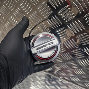 Puede incluir: Un tapón de depósito AMG plateado y negro con un anillo rojo. El tapón tiene el logotipo AMG y la palabra "OPEN". Lo sostiene una mano enguantada. El fondo es un patrón de placa de diamante.