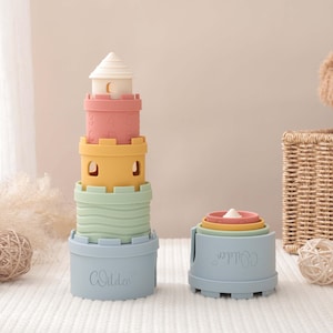 Puede incluir: Dos juegos de juguetes apilables de colores. Uno es una torre con forma de castillo, con una parte superior blanca, secciones rosas, amarillas y verdes. El otro es un conjunto de tazas anidadas. Ambos conjuntos son de color azul claro y tienen la palabra "Wilder".