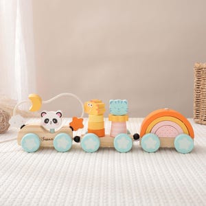 Puede incluir: Tren de juguete de madera con bloques con forma de panda, tigre y gato, y un arco iris. El tren tiene ruedas azul claro con detalles de estrellas. El tren está hecho de madera natural y pintado en colores pastel.