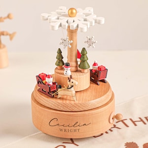 Könnte beinhalten: Eine hölzerne Weihnachtsmusikbox mit einem Schneeflocken-Design. Die Spieluhr zeigt Miniaturfiguren von Santa in einem Schlitten, einem Schneemann und Weihnachtsbäumen. Der Name "Cecilia Wright" ist auf der Basis eingraviert.