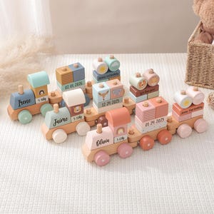 May include: A selection of pastel-coloured wooden toy trains. Each carriage displays a name and date. The trains are on a white surface. A teddy bear and basket are in the background. The trains are designed for children.