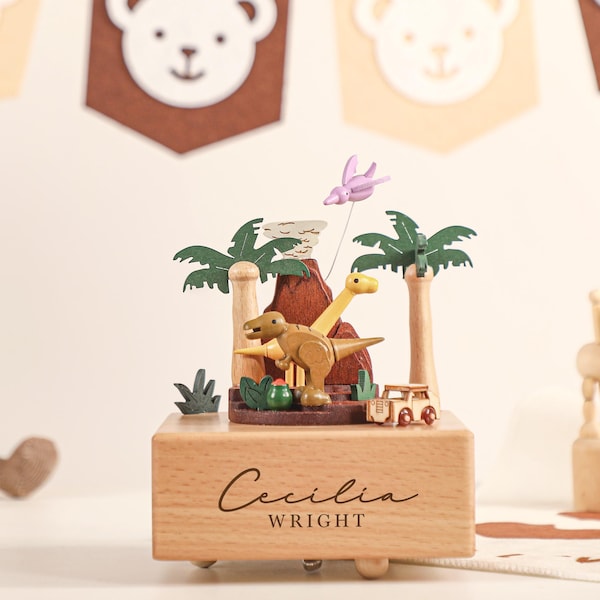 May include: Wooden music box featuring a dinosaur theme with a volcano, palm trees, and a small car. The box is engraved with the name "Cecilia Wright". The scene includes a purple pterodactyl and a yellow dinosaur. The box sits on a white surface.