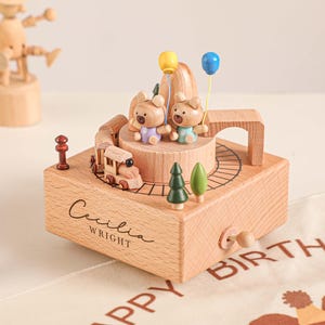 May include: A wooden music box with a train, two bear figurines holding balloons, and two small trees. The box is engraved with the name "Cecilia Wright". A wooden toy and a basket with a figurine are in the background.