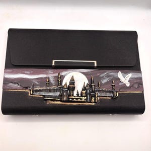 Puede incluir: Clutch negra con cierre plateado. El frente presenta un diseño detallado y en relieve de un castillo bajo la luna llena, con un pájaro blanco en vuelo. El diseño del castillo tiene detalles dorados y plateados.