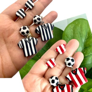 May include: Two pairs of dangle earrings featuring a football charm and a striped jersey charm. One pair has a black and white jersey charm and the other pair has a red and white jersey charm.