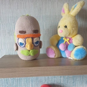 May include: Three crocheted stuffed toys on a shelf. The first toy is a brown and green character with a mustache and glasses. The second toy is a yellow bunny with pink and blue accents. The third toy is a red and orange clown with a white face.