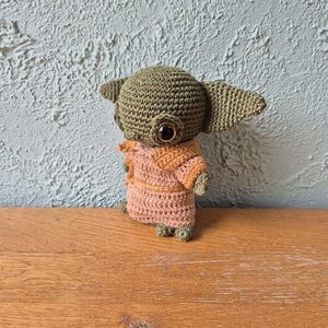 Op de afbeelding: Handgemaakte gehaakte pop van een personage met grote oren en grote ogen. De pop is voornamelijk olijfgroen, met een bruin en oranje outfit. De pop staat op een houten oppervlak, met een getextureerde, lichtgroene muur op de achtergrond.