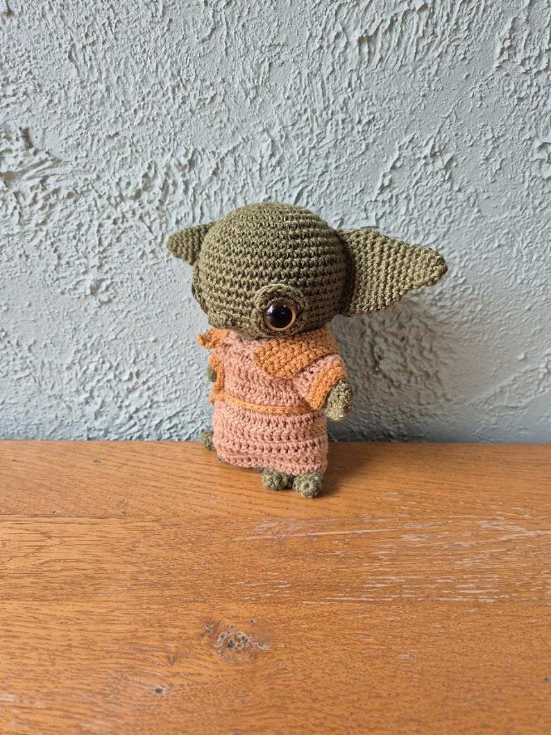 Op de afbeelding: Handgemaakte gehaakte pop van een personage met grote oren en grote ogen. De pop is voornamelijk olijfgroen, met een bruin en oranje outfit. De pop staat op een houten oppervlak, met een getextureerde, lichtgroene muur op de achtergrond.