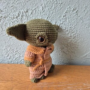 Op de afbeelding: Een handgemaakte gehaakte figuur van een personage met grote oren en grote ogen, gekleed in een bruine en beige jurk. De figuur is gemaakt van groen en beige garen en staat op een houten oppervlak.