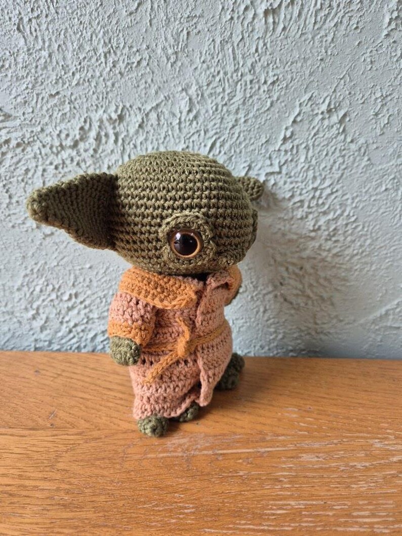 Op de afbeelding: Een handgemaakte gehaakte figuur van een personage met grote oren en grote ogen, gekleed in een bruine en beige jurk. De figuur is gemaakt van groen en beige garen en staat op een houten oppervlak.