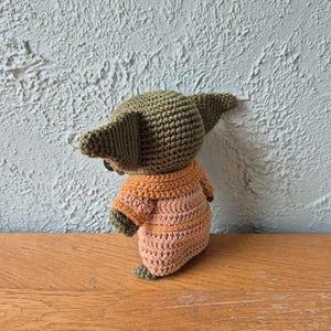 Op de afbeelding: Een handgemaakte gehaakte pop van een personage met grote oren, gekleed in een beige en oranje outfit. De pop is gemaakt met een strak steekpatroon en de details omvatten kleine voetjes en een ronde kop. De pop wordt gepresenteerd op een houten oppervlak.
