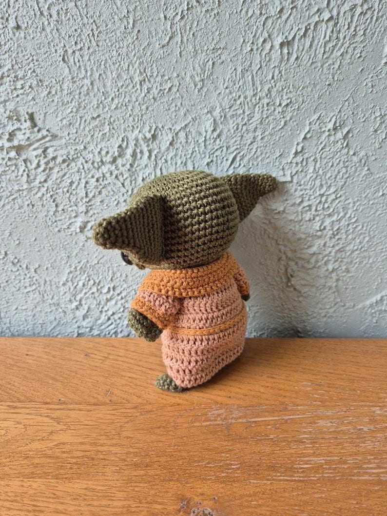 Op de afbeelding: Een handgemaakte gehaakte pop van een personage met grote oren, gekleed in een beige en oranje outfit. De pop is gemaakt met een strak steekpatroon en de details omvatten kleine voetjes en een ronde kop. De pop wordt gepresenteerd op een houten oppervlak.